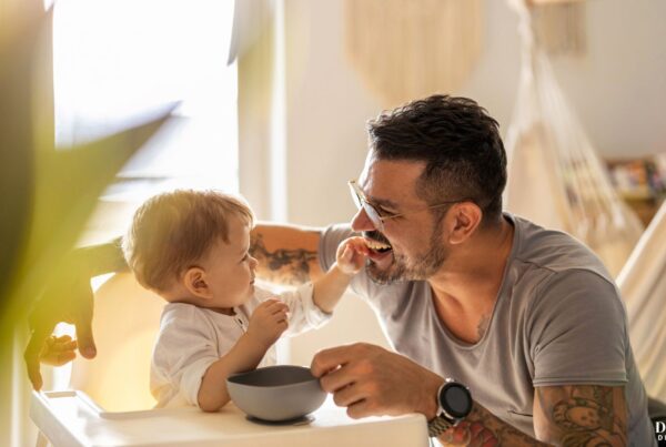 A father feeding and playing with his child at the kitchen table.