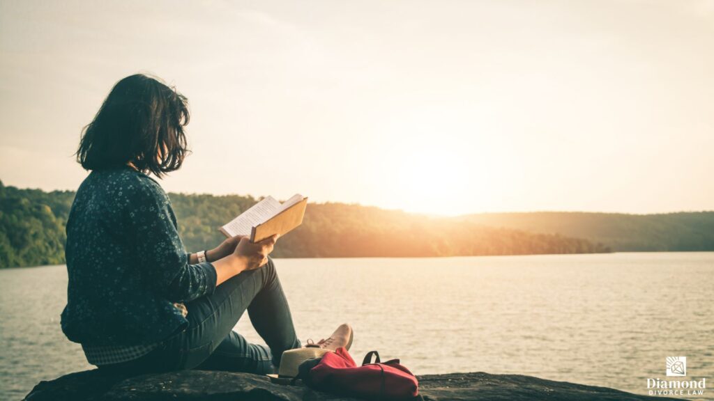 A woman sitting by a lake reading a book.