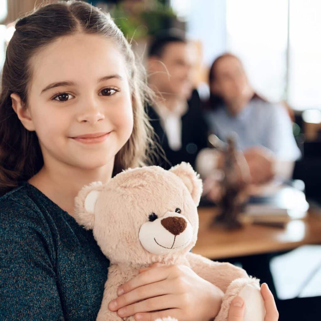 A little girl holding a teddy bear with a couple sitting at a table behind her.